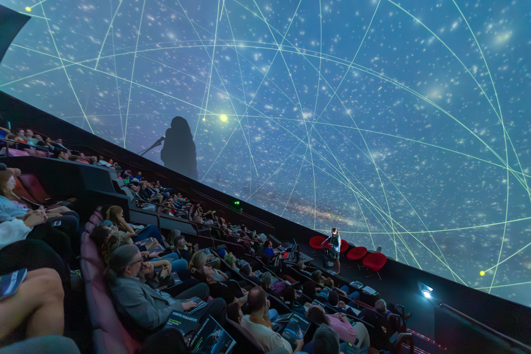 Speaker presenting beneath a large planetarium dome filled with projected stars and orbital tracks